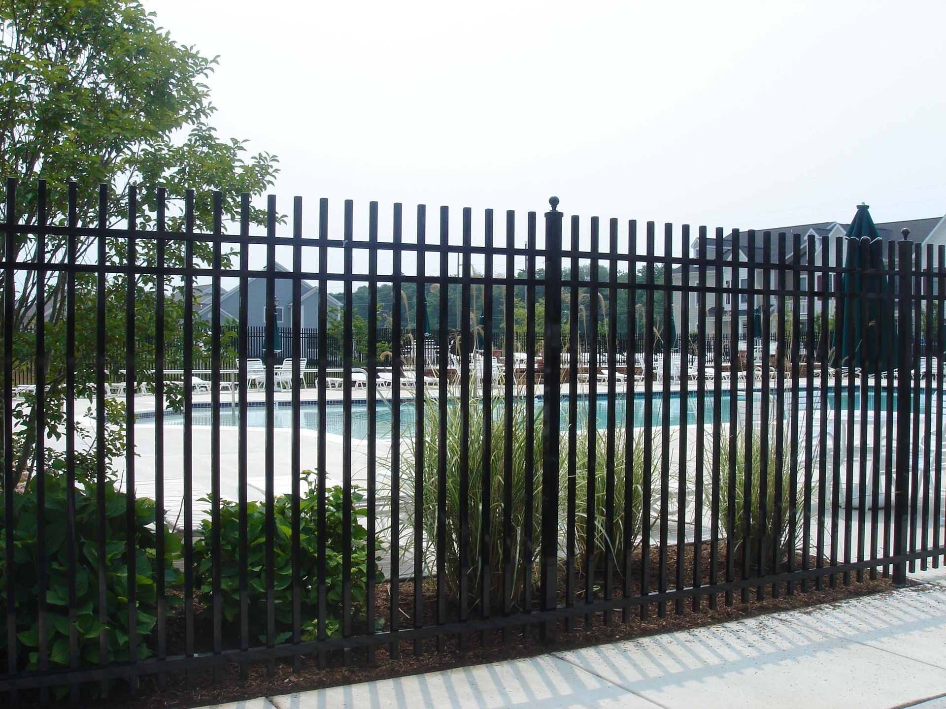 Black metal fence surrounding a swimming pool with a glimpse of the pool and buildings beyond.