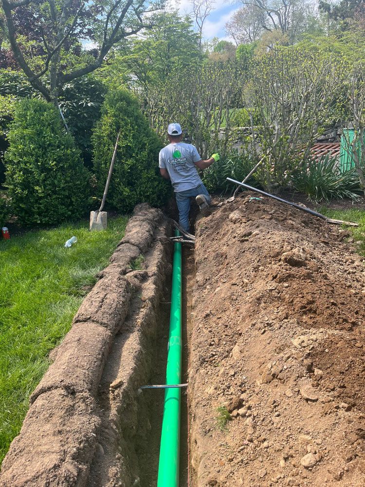 Man installing green pipe in a trench outdoors; shovel and trees visible.
