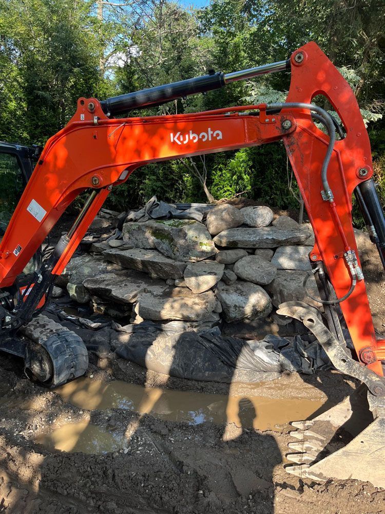 Orange Kubota excavator moving large rocks near muddy water, outdoors.