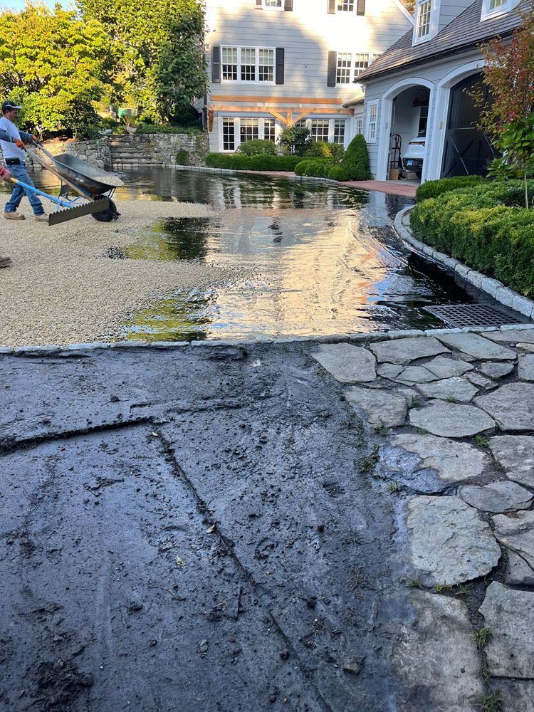 Water covers a driveway. A man uses a leaf blower. Large house in the background.