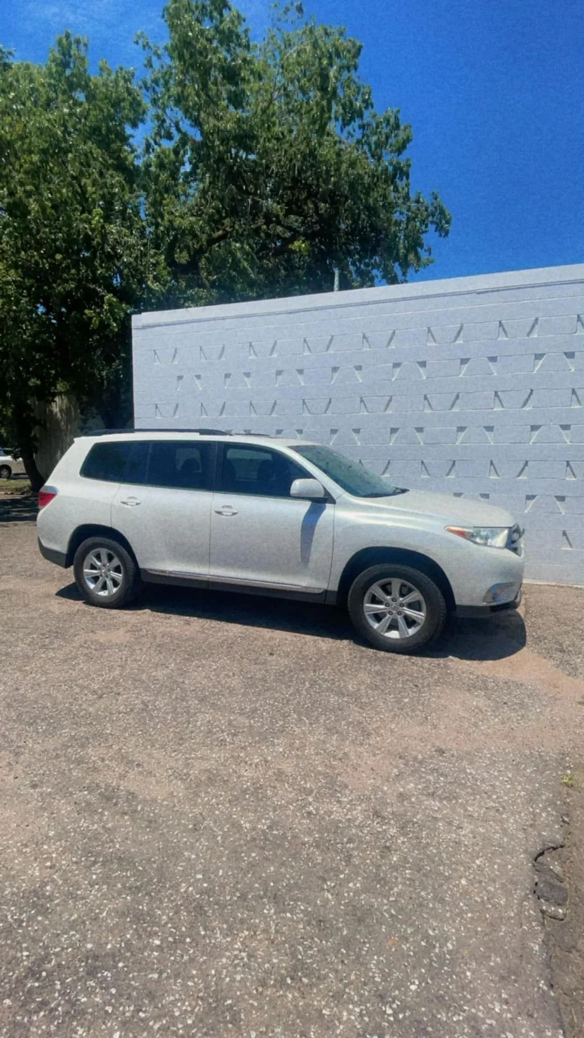 White Toyota Highlander parked next to a white building under a blue sky, near a tree.