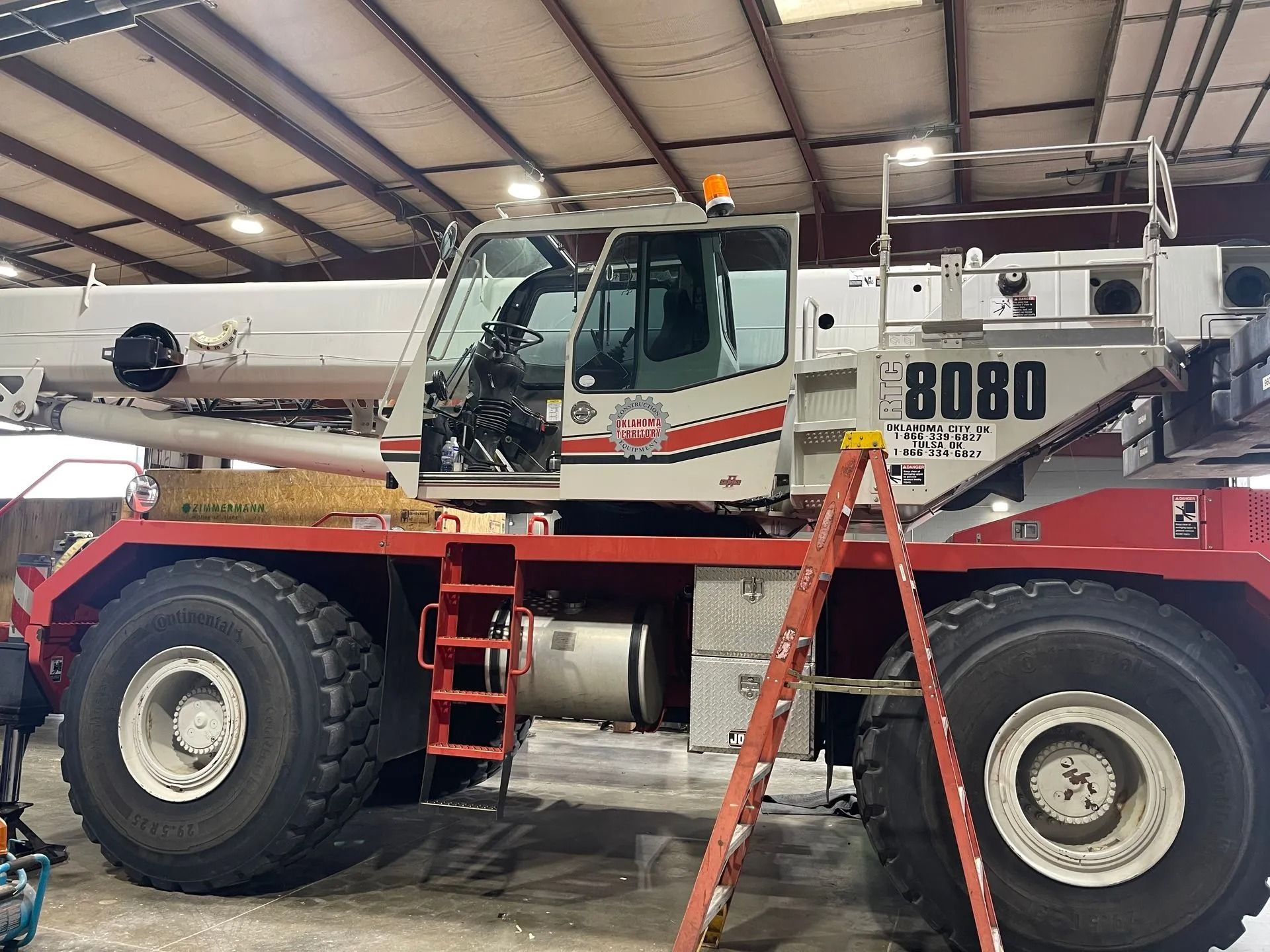 Red and white crane with large tires, parked inside a building. Ladder is leaning against it. 