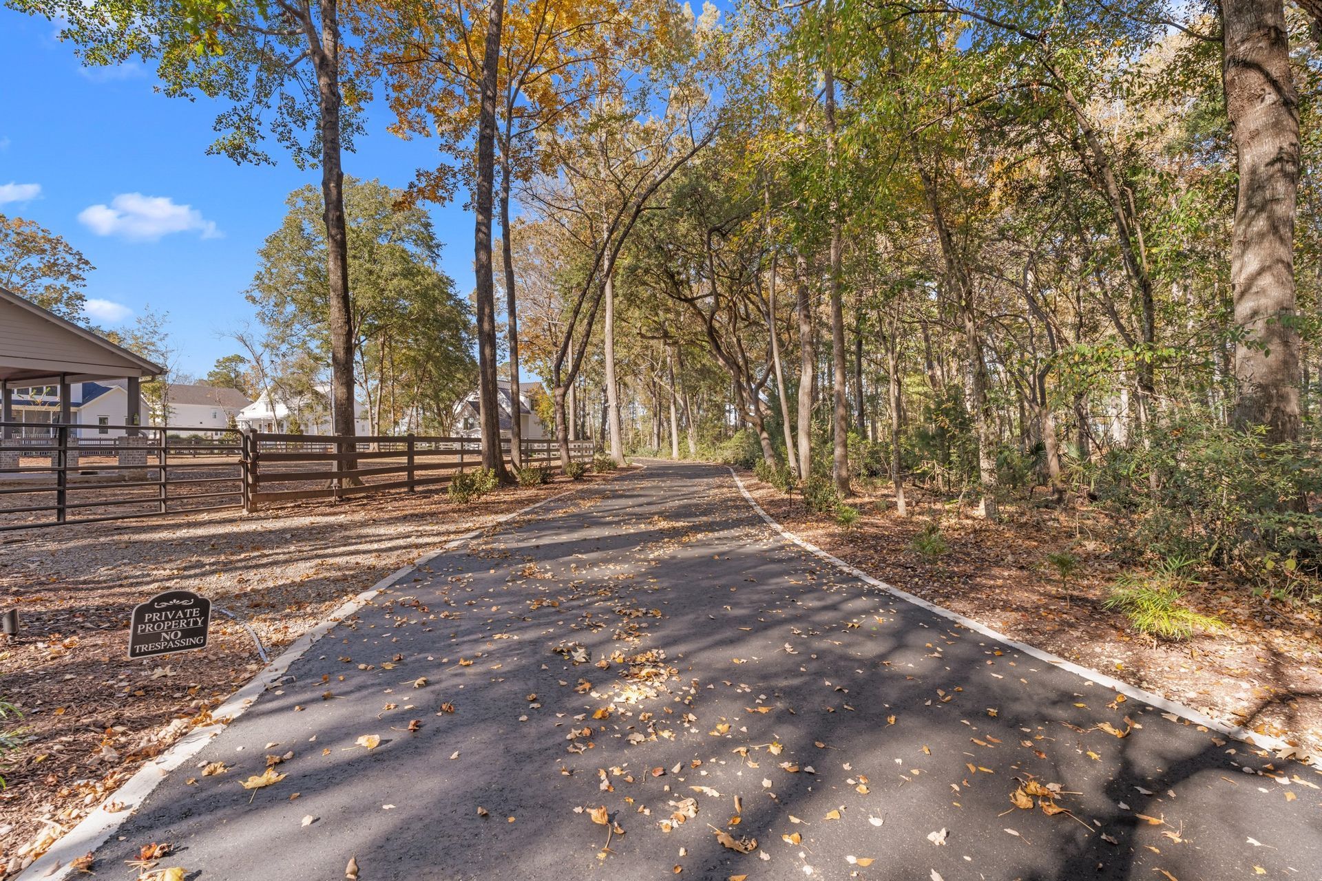 Paved road curving through a wooded area with autumn leaves and a fenced house on the left.