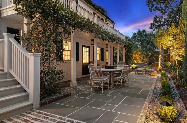 Stone patio beside a house at dusk, with outdoor dining chairs, lit windows, and garden trees.