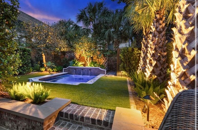Twilight backyard with lit palm trees and a small pool beside a stone patio and lawn