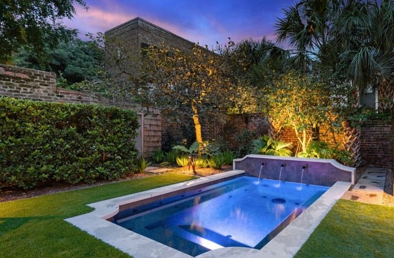 Lit backyard pool at dusk with stone walls, lush greenery, and warm landscape lighting