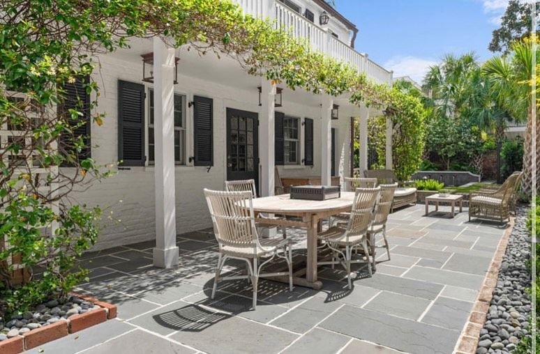 Sunlit patio with wicker dining table and chairs beside a white house and lush greenery