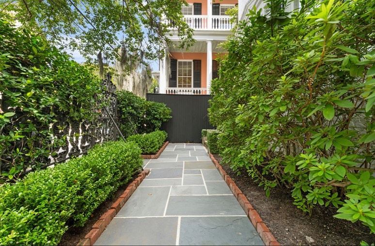 Stone walkway leading to a townhouse entrance, bordered by lush green shrubs and trees.