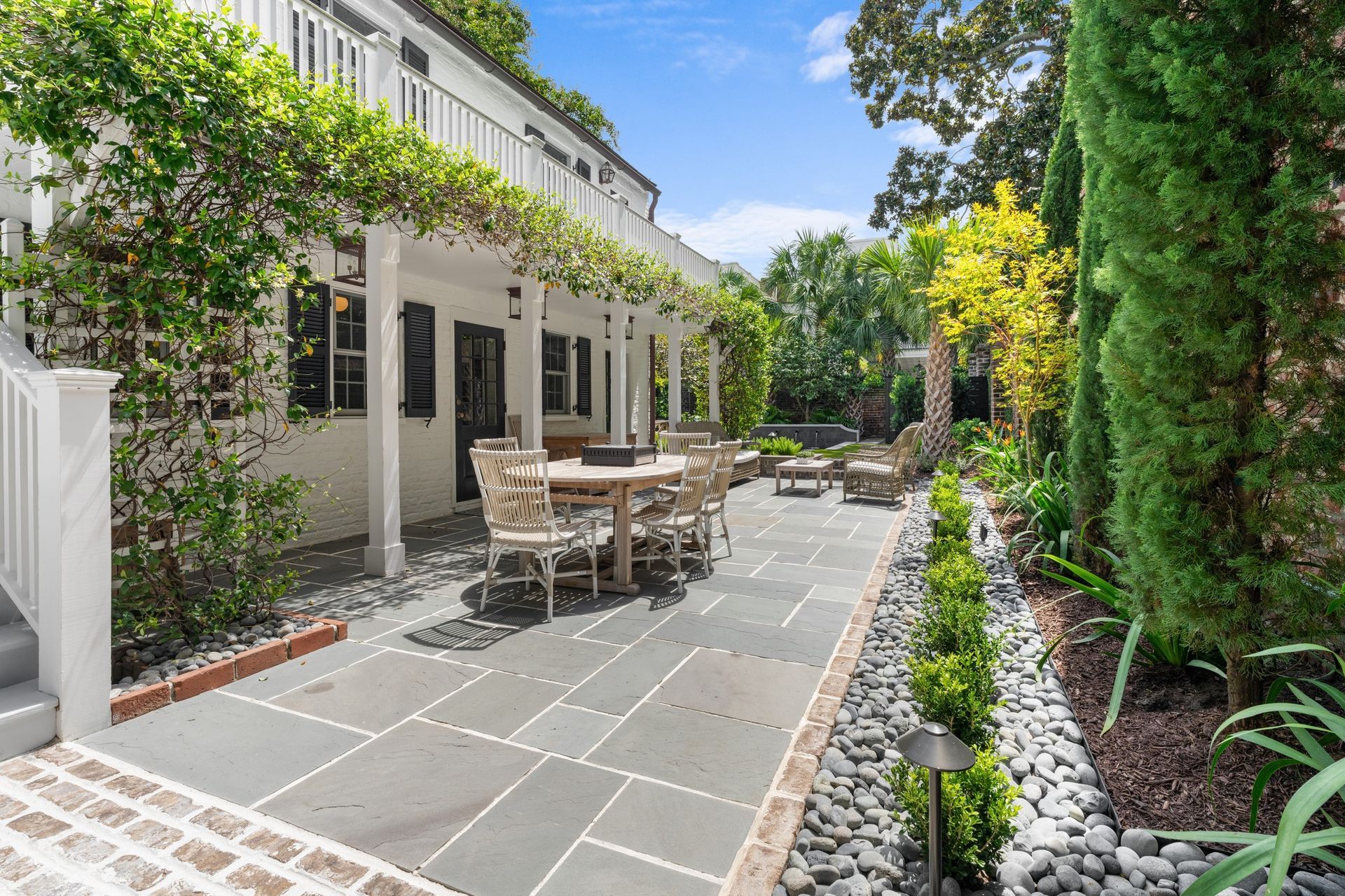 Outdoor stone patio beside a house, with dining table, chairs, and lush green landscaping on a sunny day
