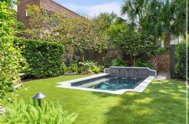 Small backyard pool surrounded by lush greenery and a stone patio in bright sunlight