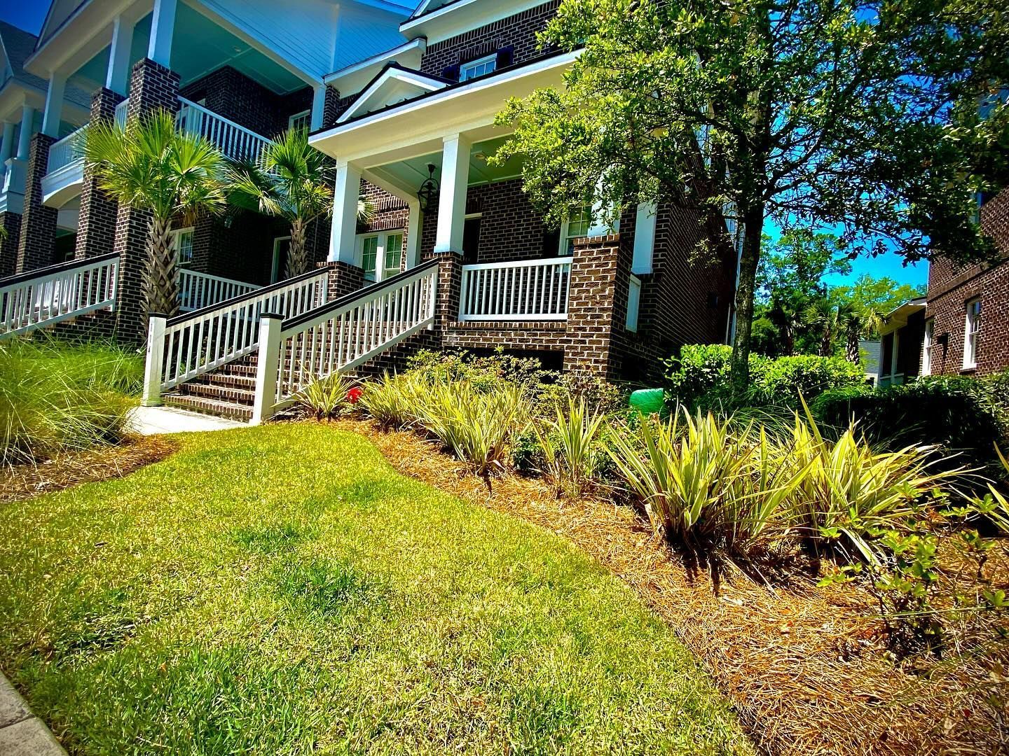 House with white porch and stairs on a sunny grassy hillside surrounded by trees and shrubs