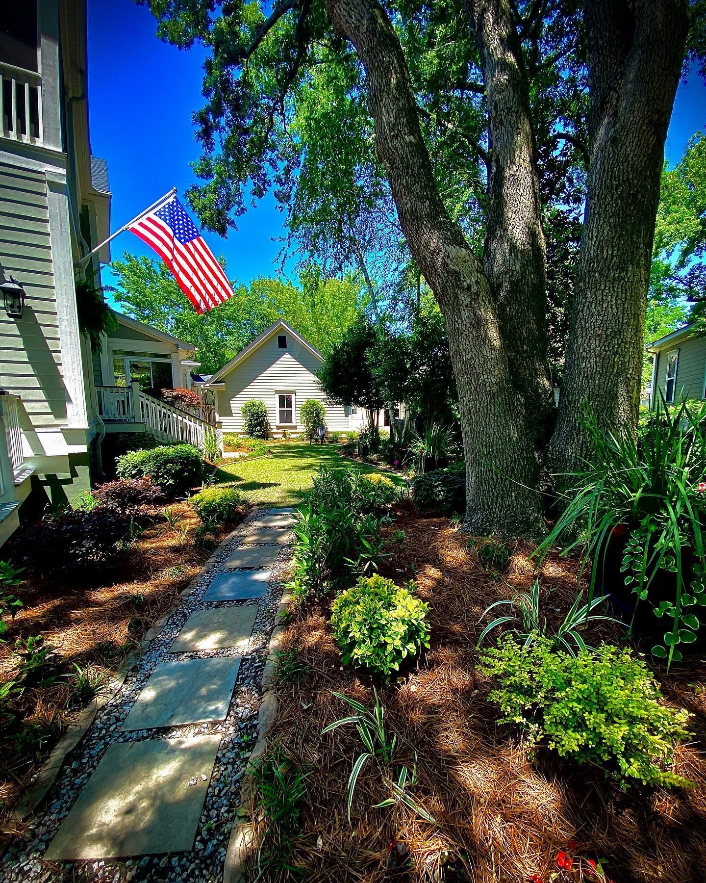 Sunlit garden path beside a house, with an American flag, trees, and a small cottage in the distance