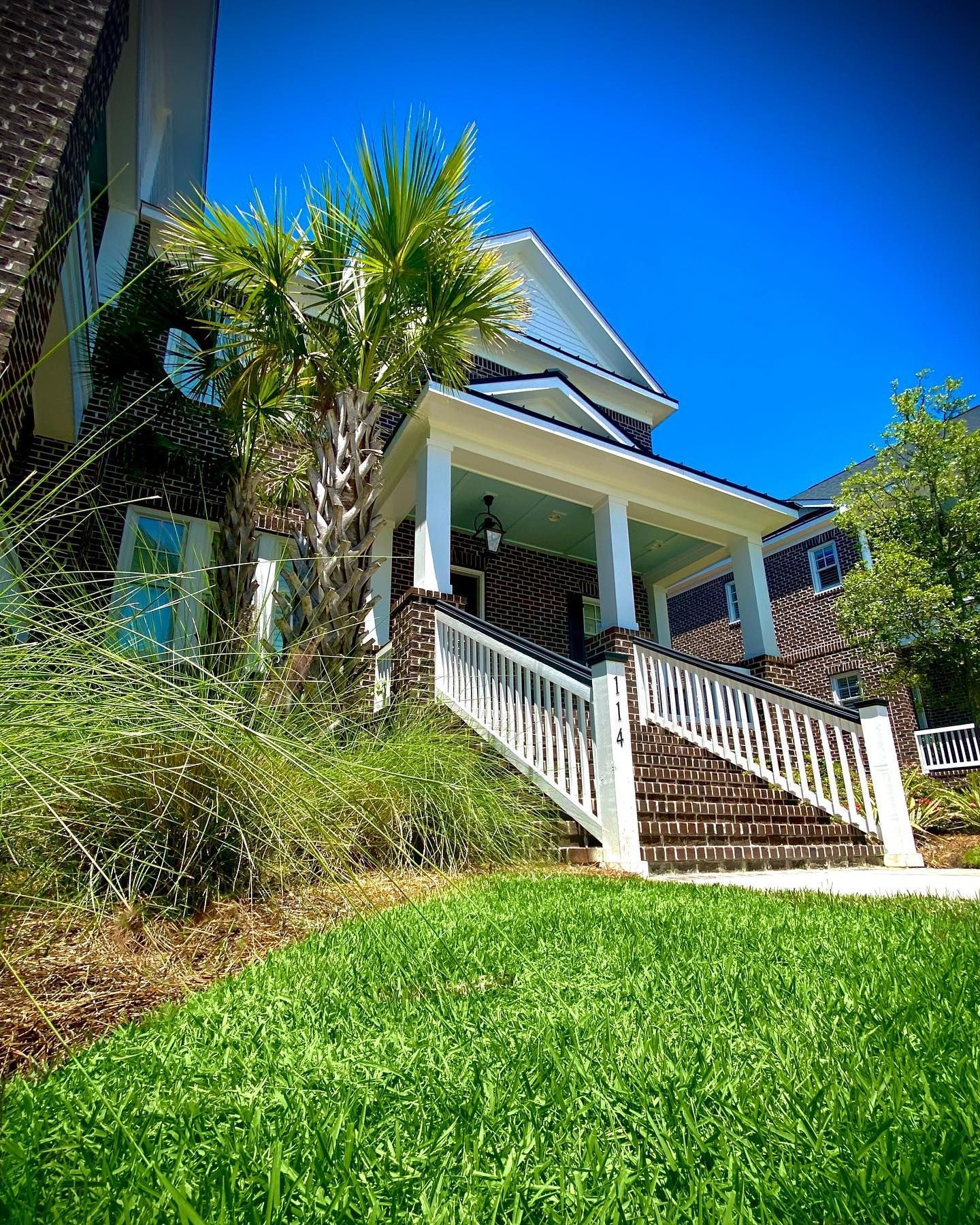Front porch of a house with white railings, stairs, palm trees, and bright green lawn under a blue sky