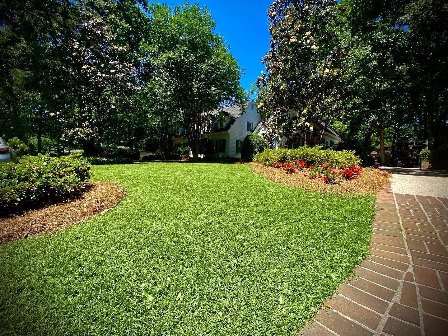 Sunny landscaped yard with green lawn, flower beds, trees, and a brick walkway leading to a house
