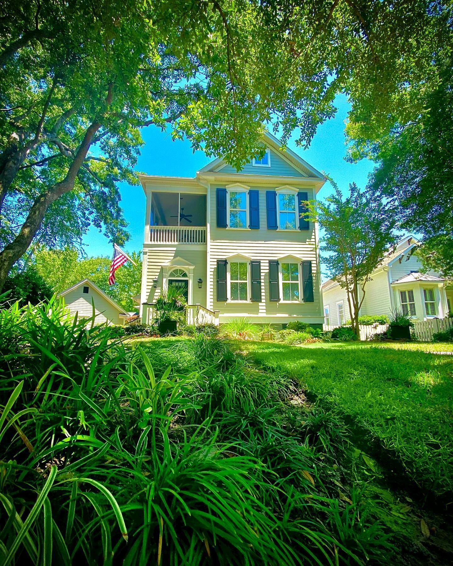 Two-story white house with green shutters, framed by trees and lush green garden under a bright blue sky