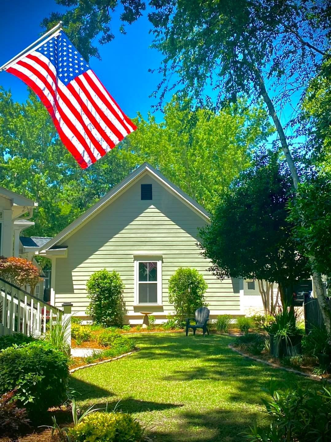 Small cream house with a red, white, and blue American flag waving in a sunny yard