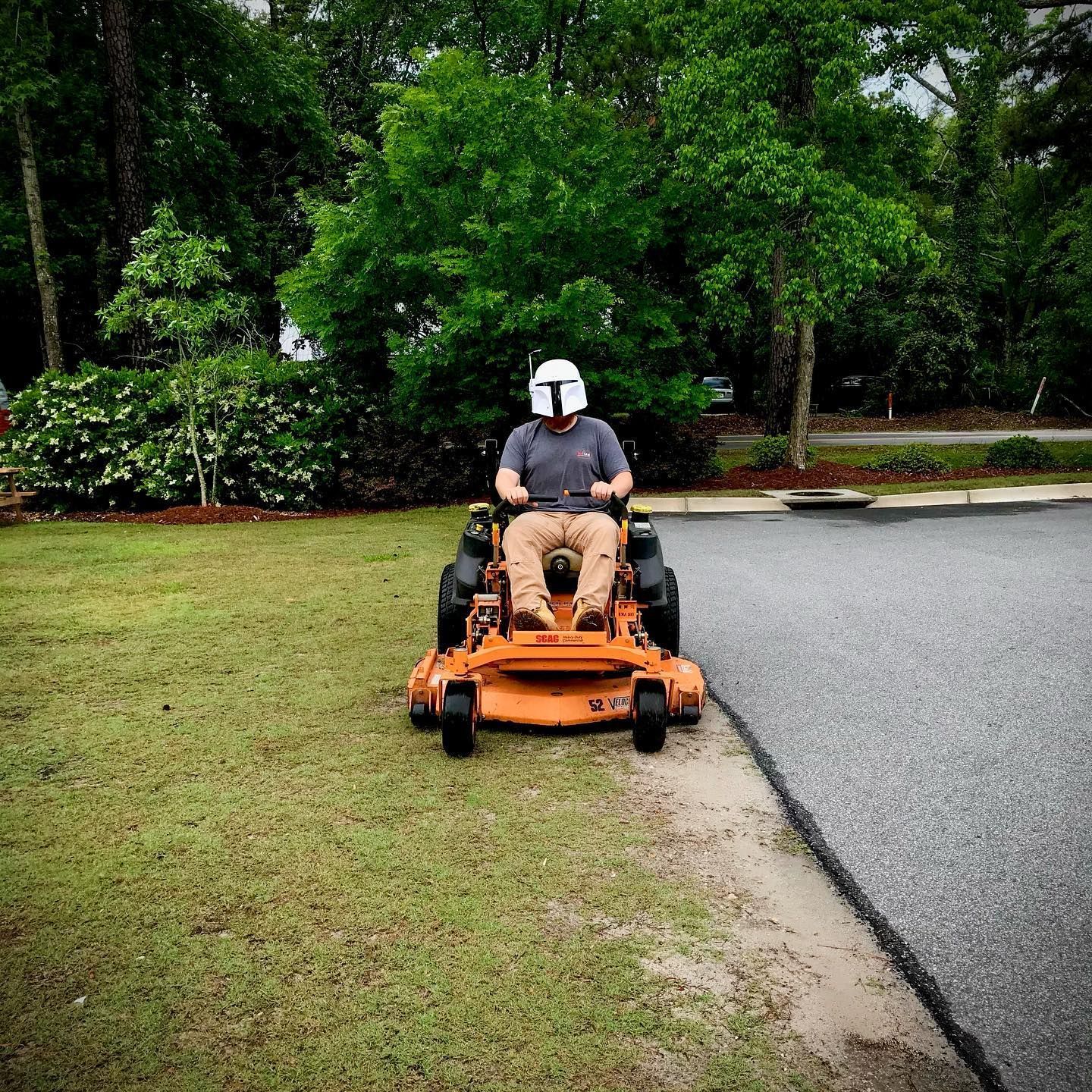 Person mowing grass beside a paved road with an orange riding mower in a green yard