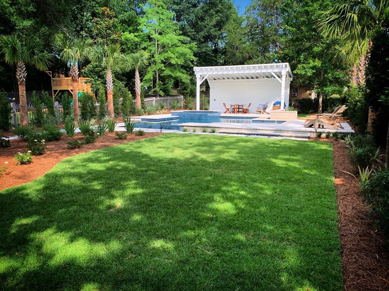 Lush backyard with green lawn, pool, and white patio pergola surrounded by trees and landscaping