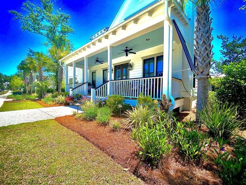 Bright porch of a white house with black railing, blue sky, palm tree, and landscaped yard