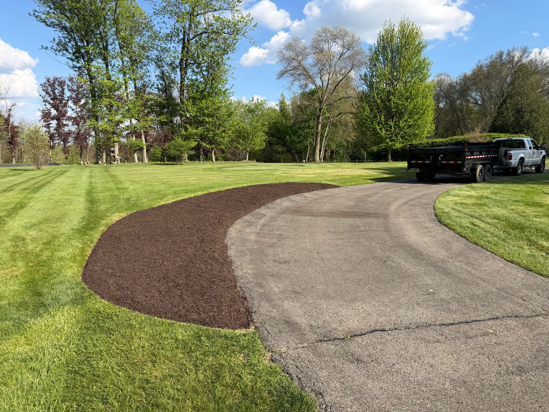 Curved driveway with fresh mulch border beside a grassy yard and trees, with a truck and trailer in the background