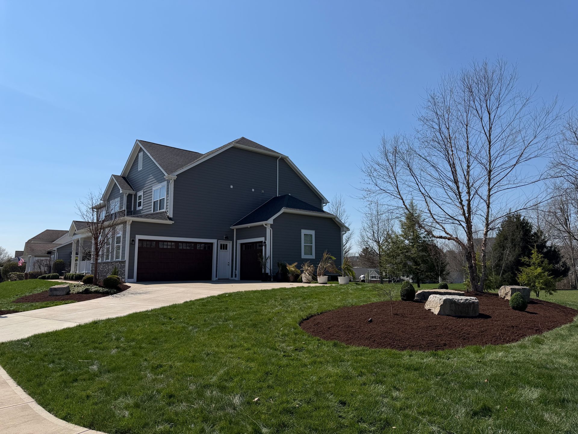 Suburban house with dark siding, two-car garage, green lawn, and landscaped front yard under a clear blue sky