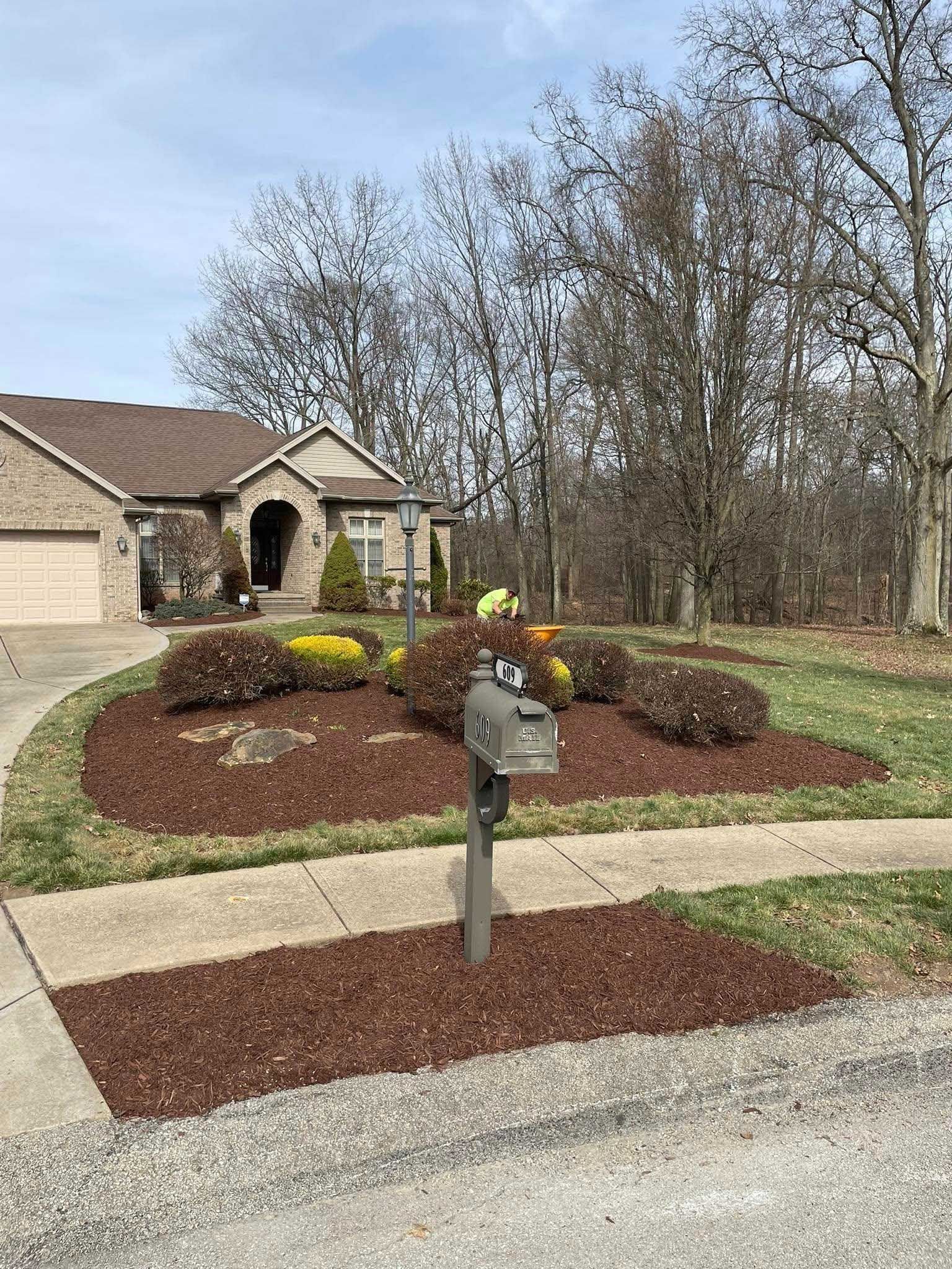 A house with a circular flower bed and a mailbox in front, all bordered with brown mulch, under a cloudy sky.