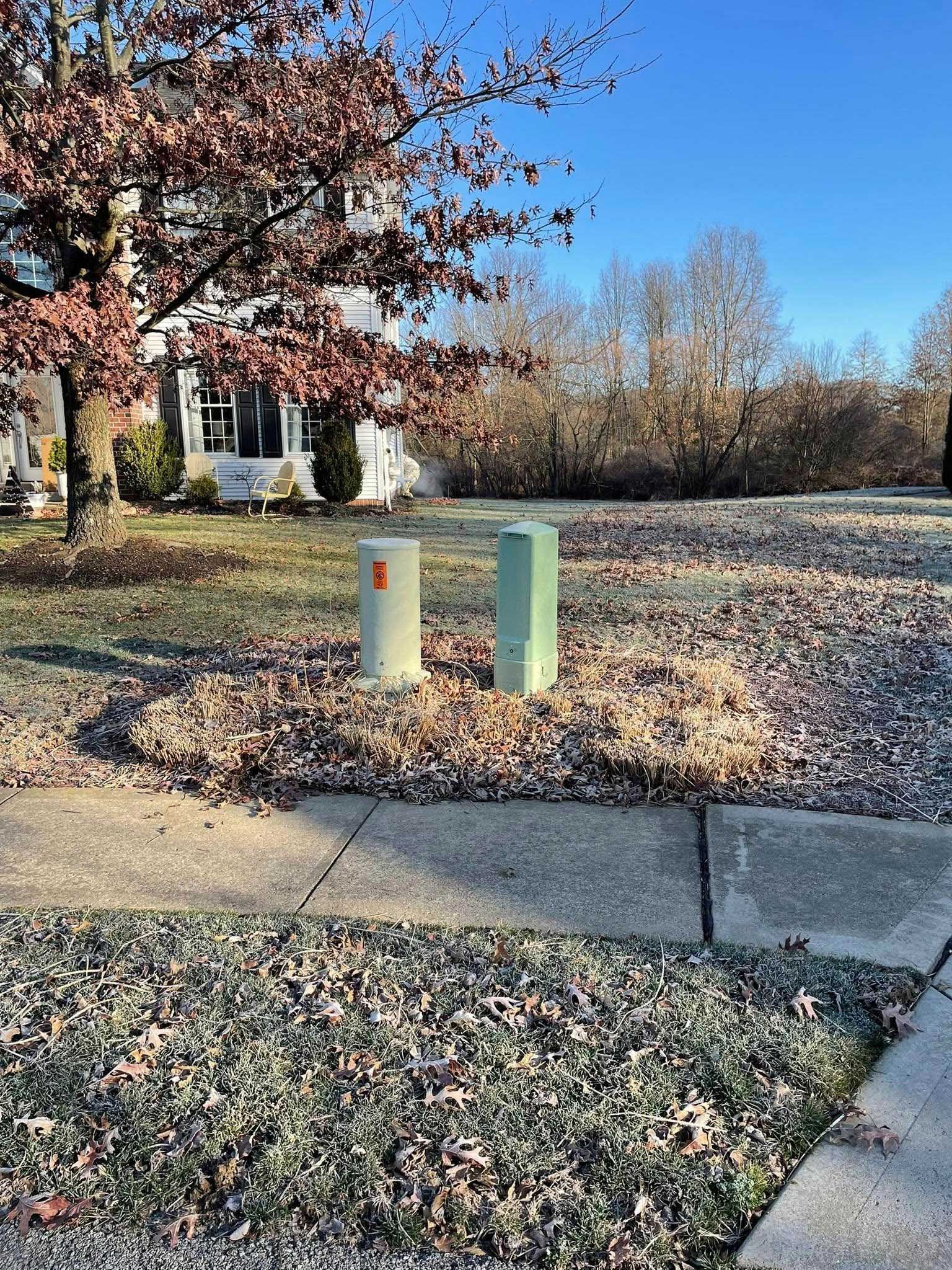 Two utility boxes sit in a yard covered with fallen leaves, with a tree and blue sky in the background.