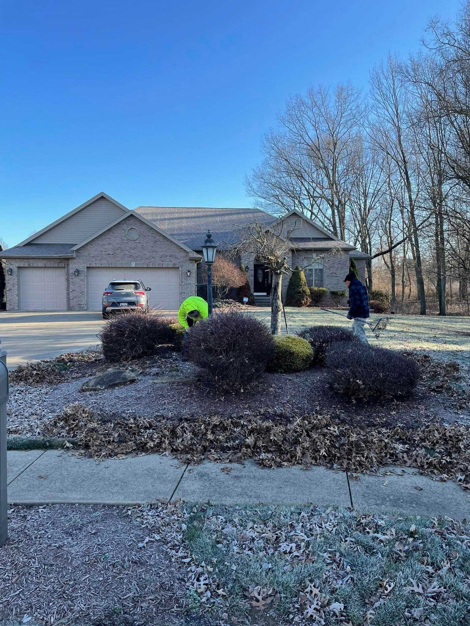 Two people raking leaves in front of a brick house with a two-car garage on a sunny day. The yard is covered in fallen leaves.