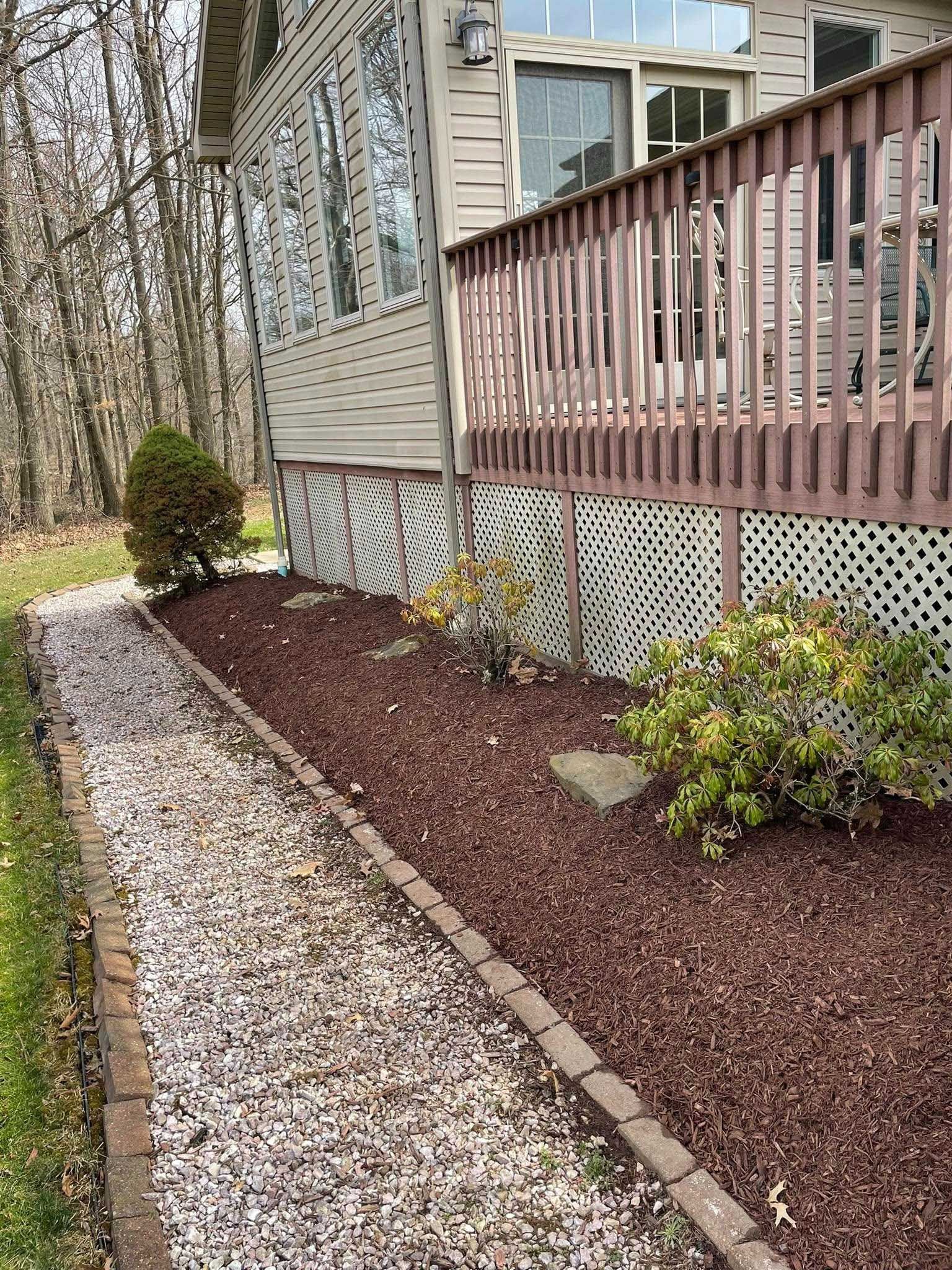 A house exterior with a wooden deck, bordered by a brick and gravel walkway. Red mulch covers the ground near the deck, with some shrubbery.