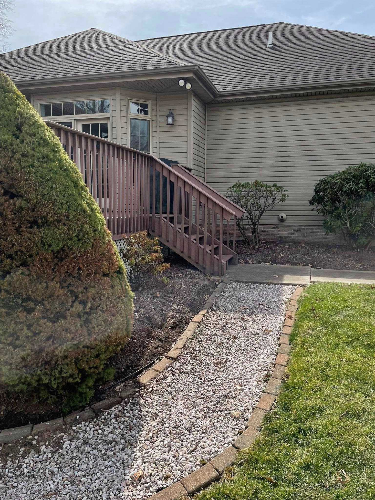 A wooden ramp and steps lead to a house entrance. A gravel path borders green lawn and bushes.