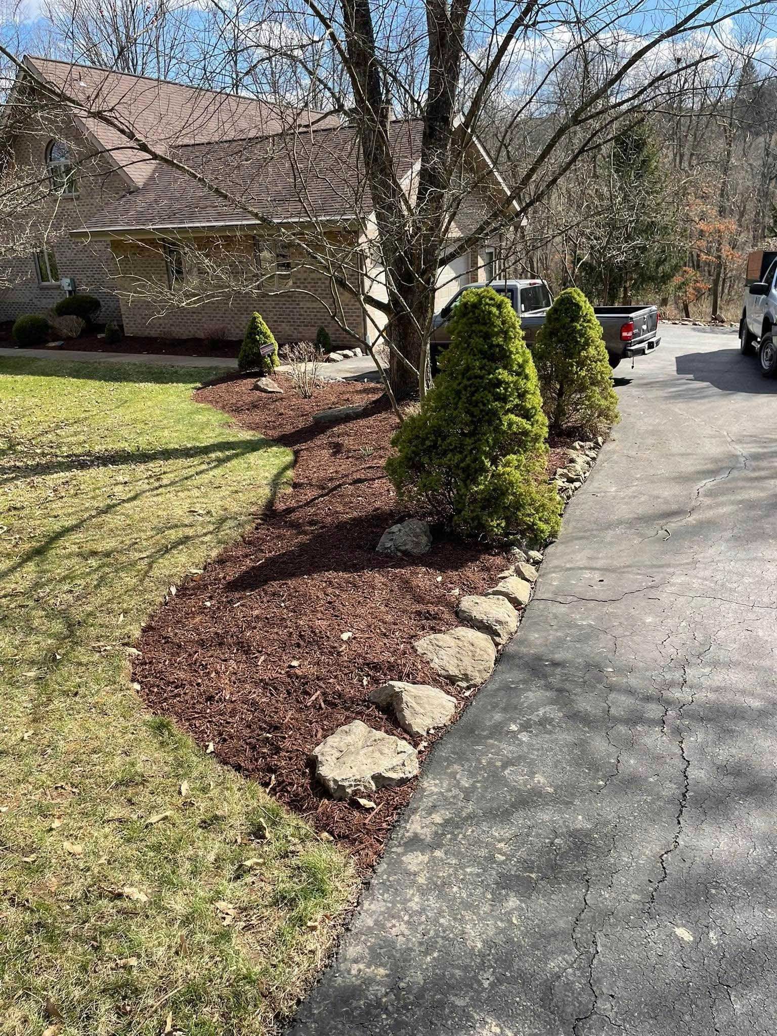 A house with a driveway, trees, and landscaped garden beds with reddish mulch.