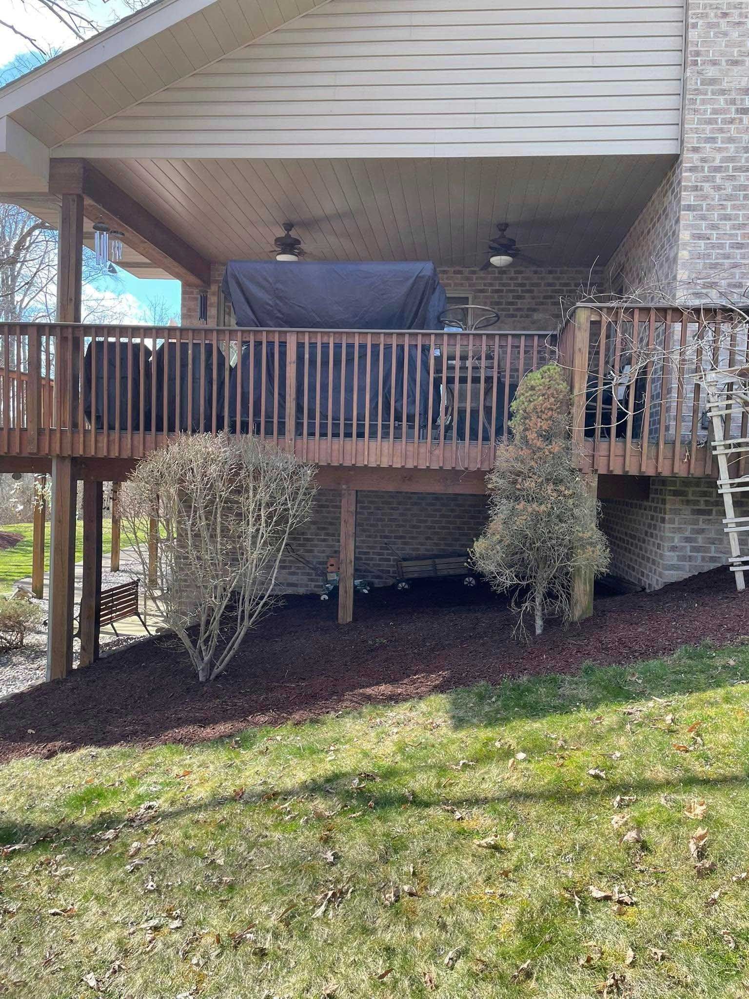 Elevated wooden deck attached to a brick house. Covered patio area with furniture, surrounded by shrubs and mulch.