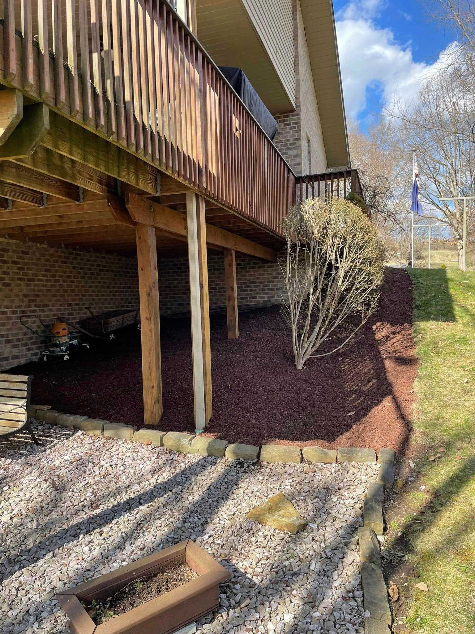Wooden deck supported by light-colored posts; landscaping with red mulch, gravel, and a small shrub. A building and a blue sky are in the background.