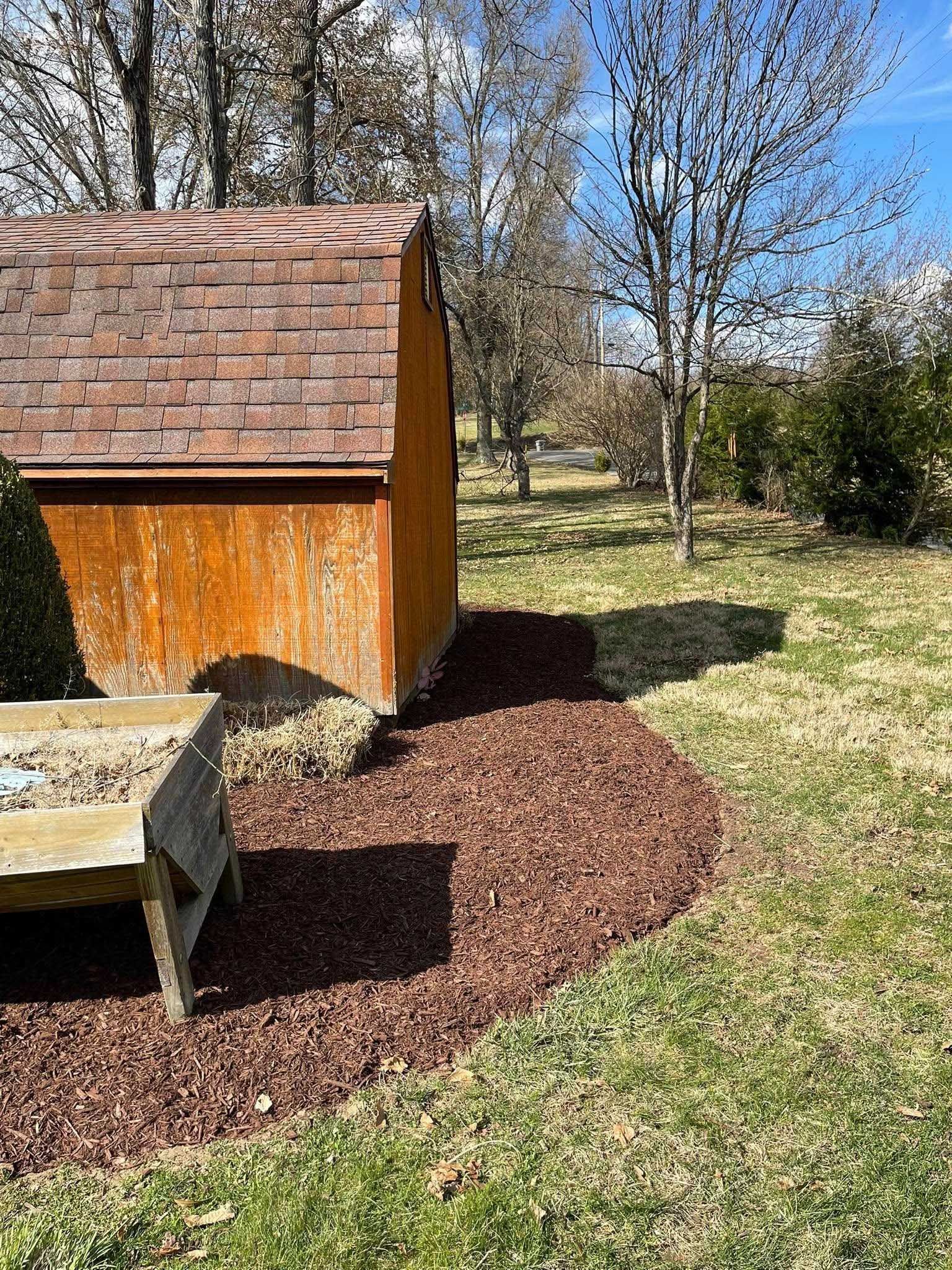 A wooden shed with a brown roof and a stone bench sit next to a bed of brown mulch on a sunny lawn.