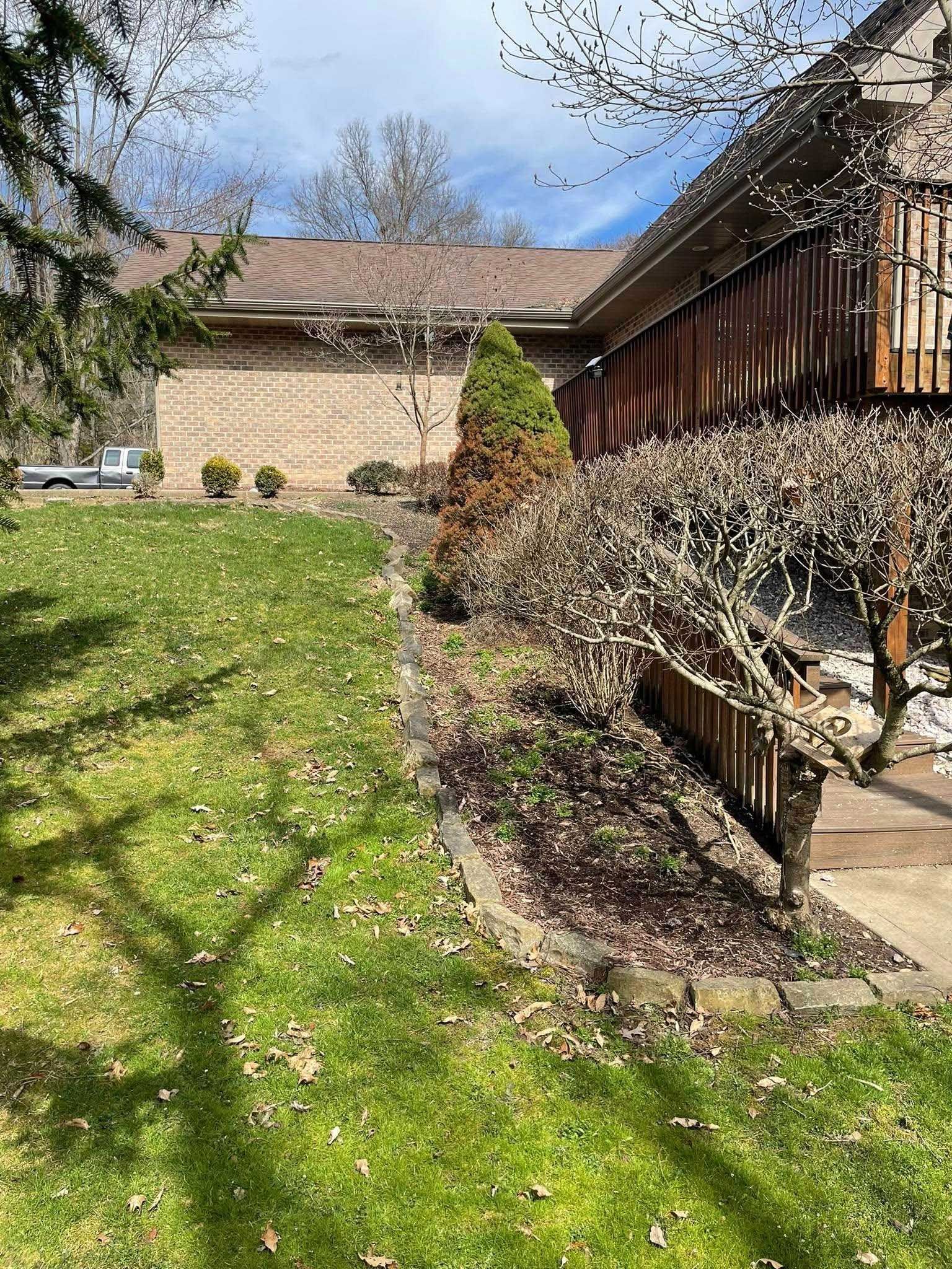 Lawn with green grass slopes up to a brick house with a wooden deck. Trees and shrubs are in the yard.