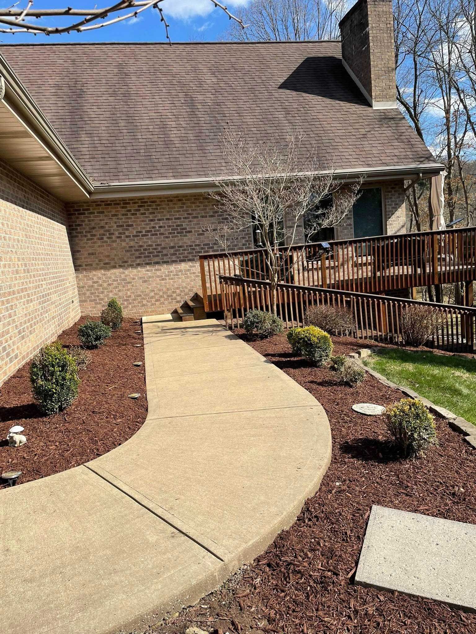 A concrete pathway leads to a house with a wooden deck and ramp. Landscaping includes mulch, shrubs, and a brick exterior.