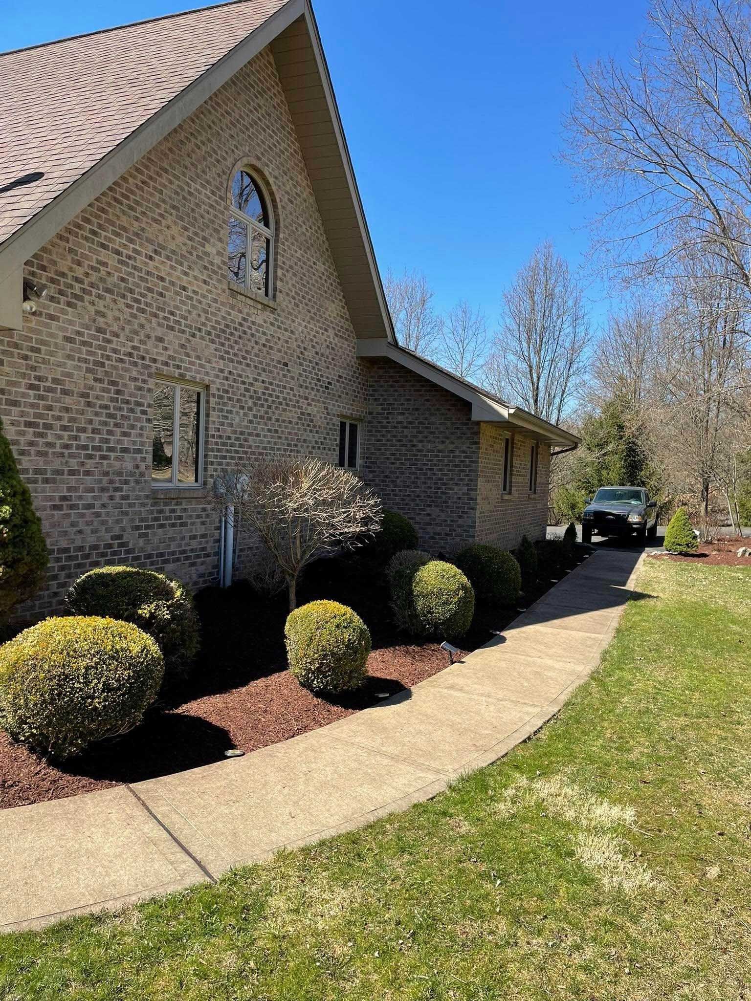 Side view of a brick house with a curving concrete walkway lined with bushes and mulch, with a car parked in the driveway.
