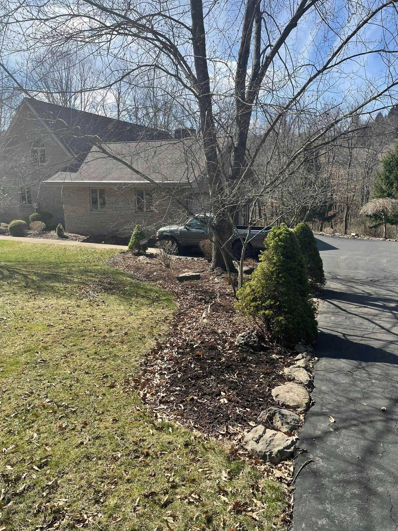 A house with brown bricks and a dark car parked in front, surrounded by a tree and bushes. Fallen brown leaves cover the ground.