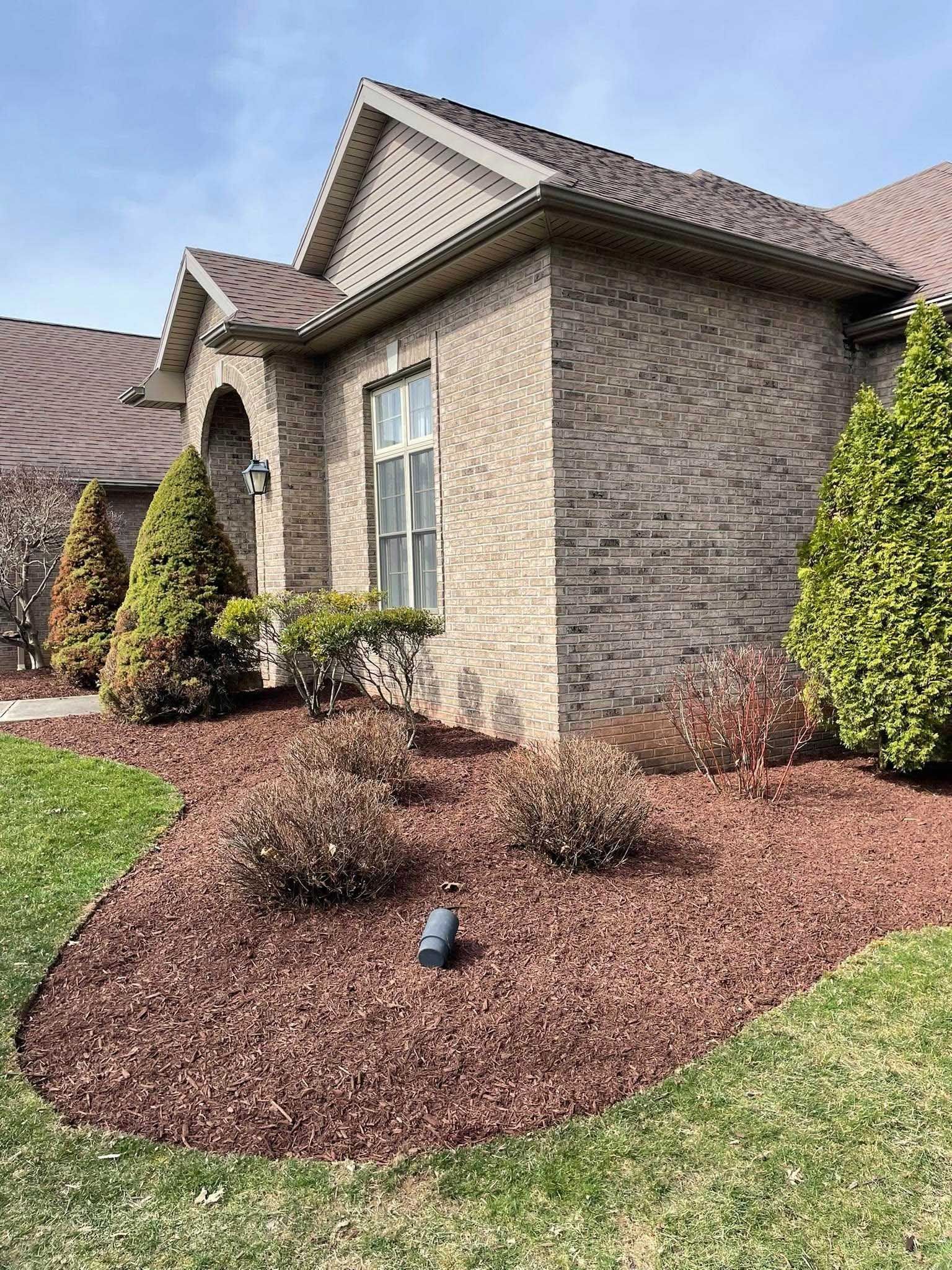 A brick house with brown mulch and landscaping. Green grass surrounds the flower beds with small bushes.