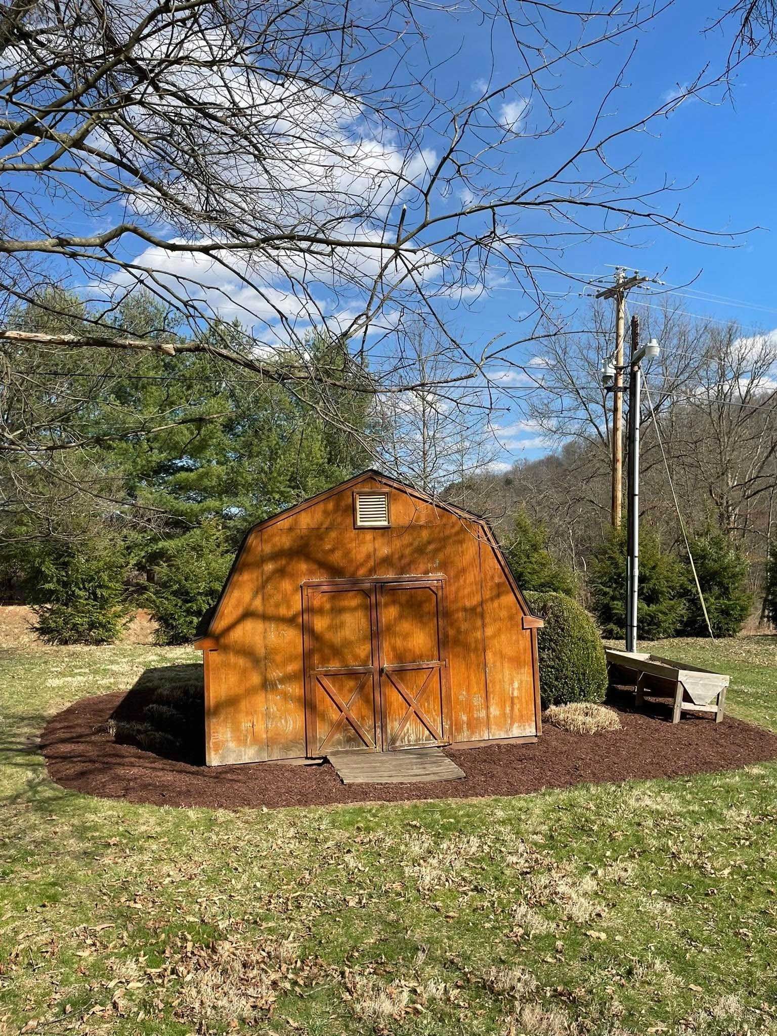 Wooden shed in a grassy yard, surrounded by mulch and trees. A utility pole stands to the right. Blue sky visible above.