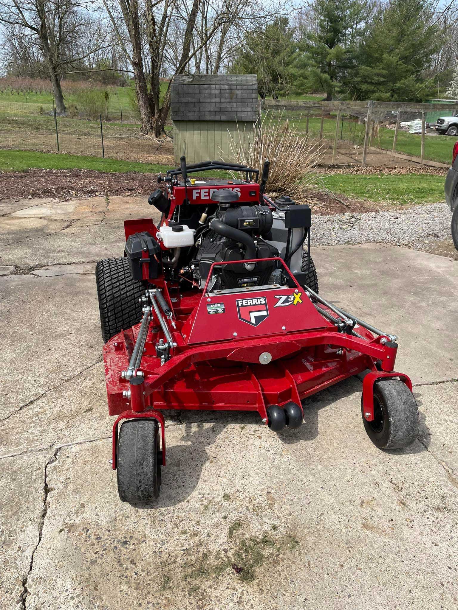 Red zero-turn lawnmower on a concrete surface with a shed and trees in the background.
