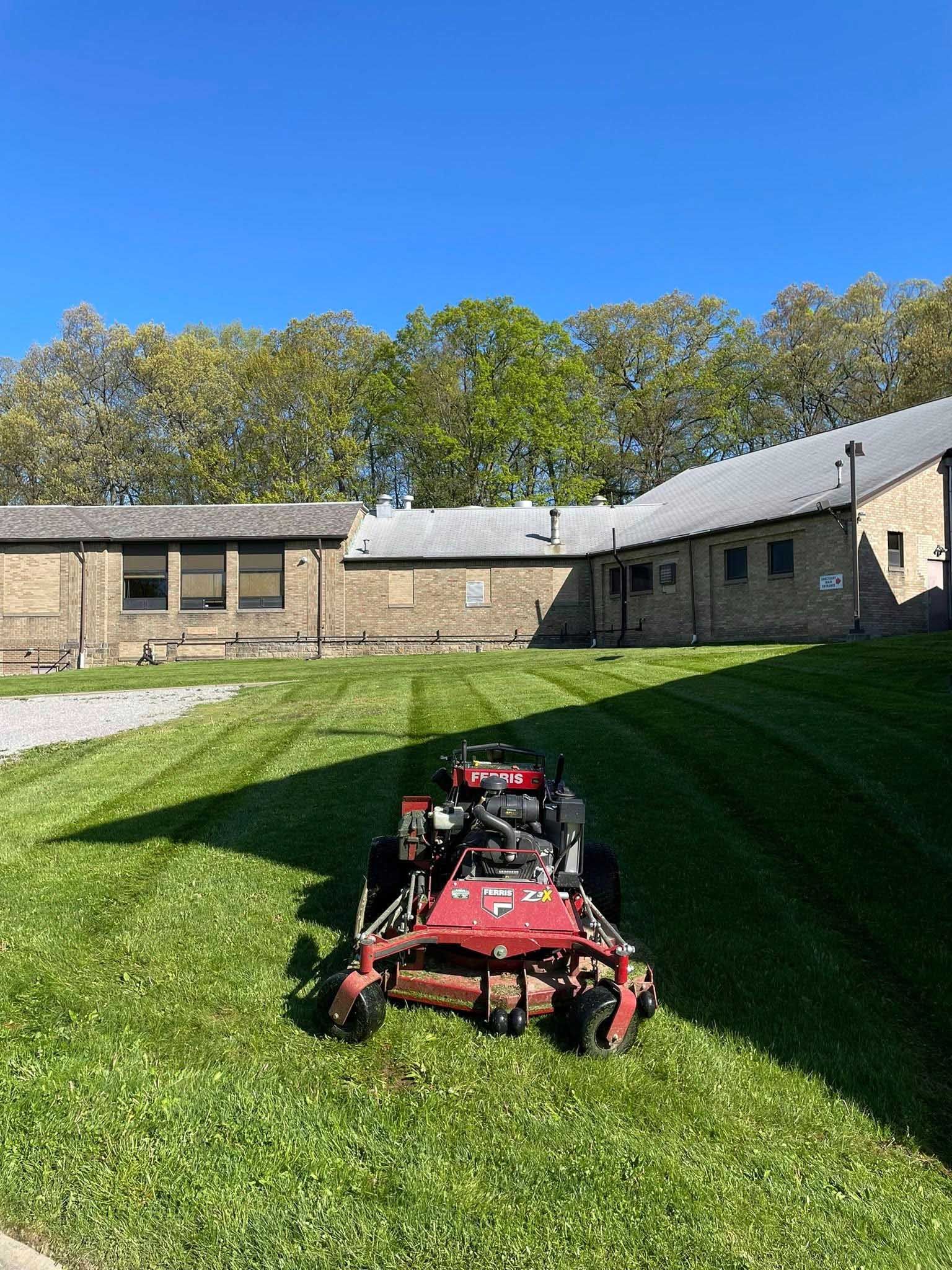 Lawnmower cutting grass on a sunny day in front of a brick building with trees in the background.