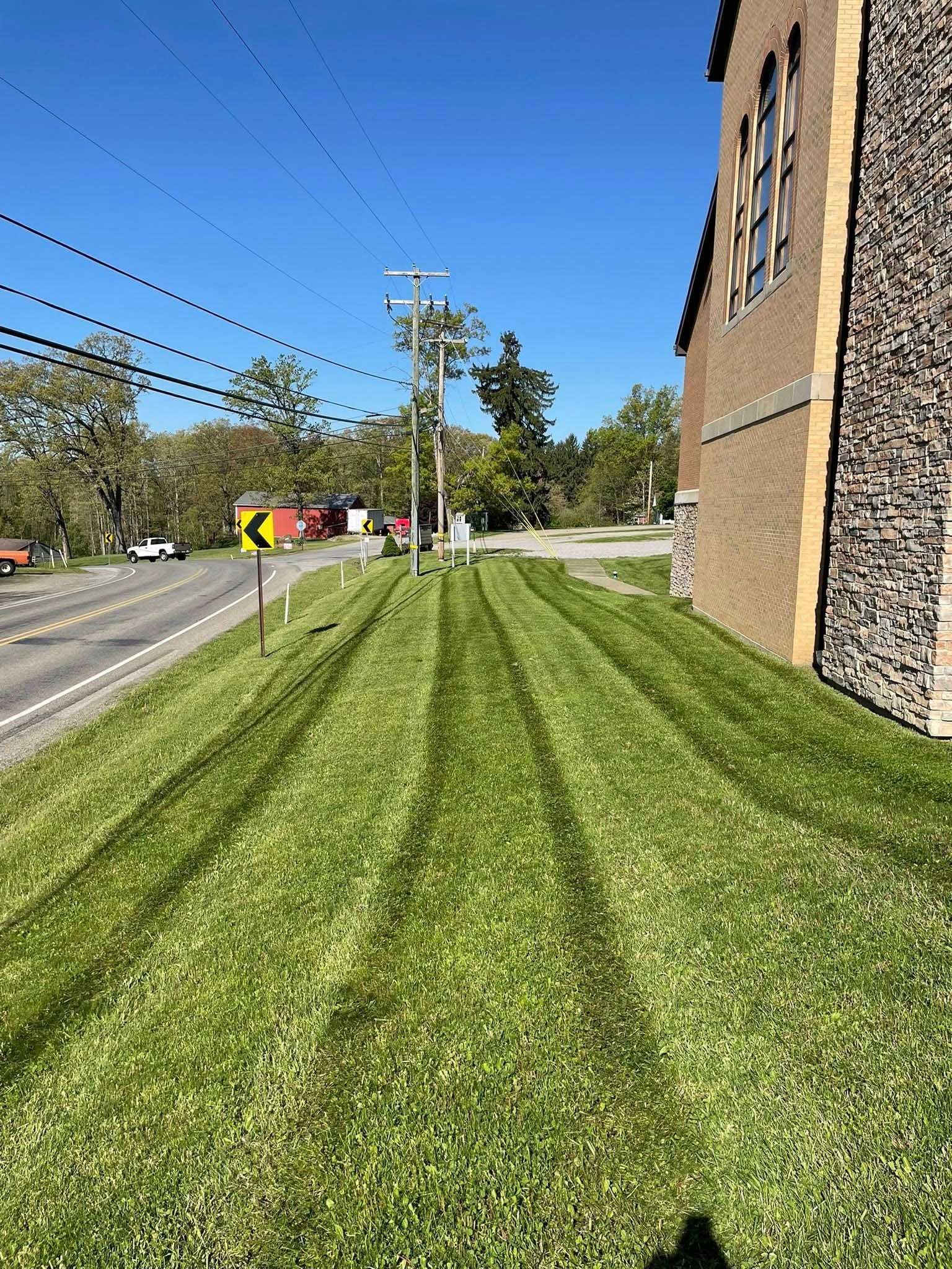 Lawn with mowing stripes next to a brick building and road on a sunny day. A power pole and a sign are visible.