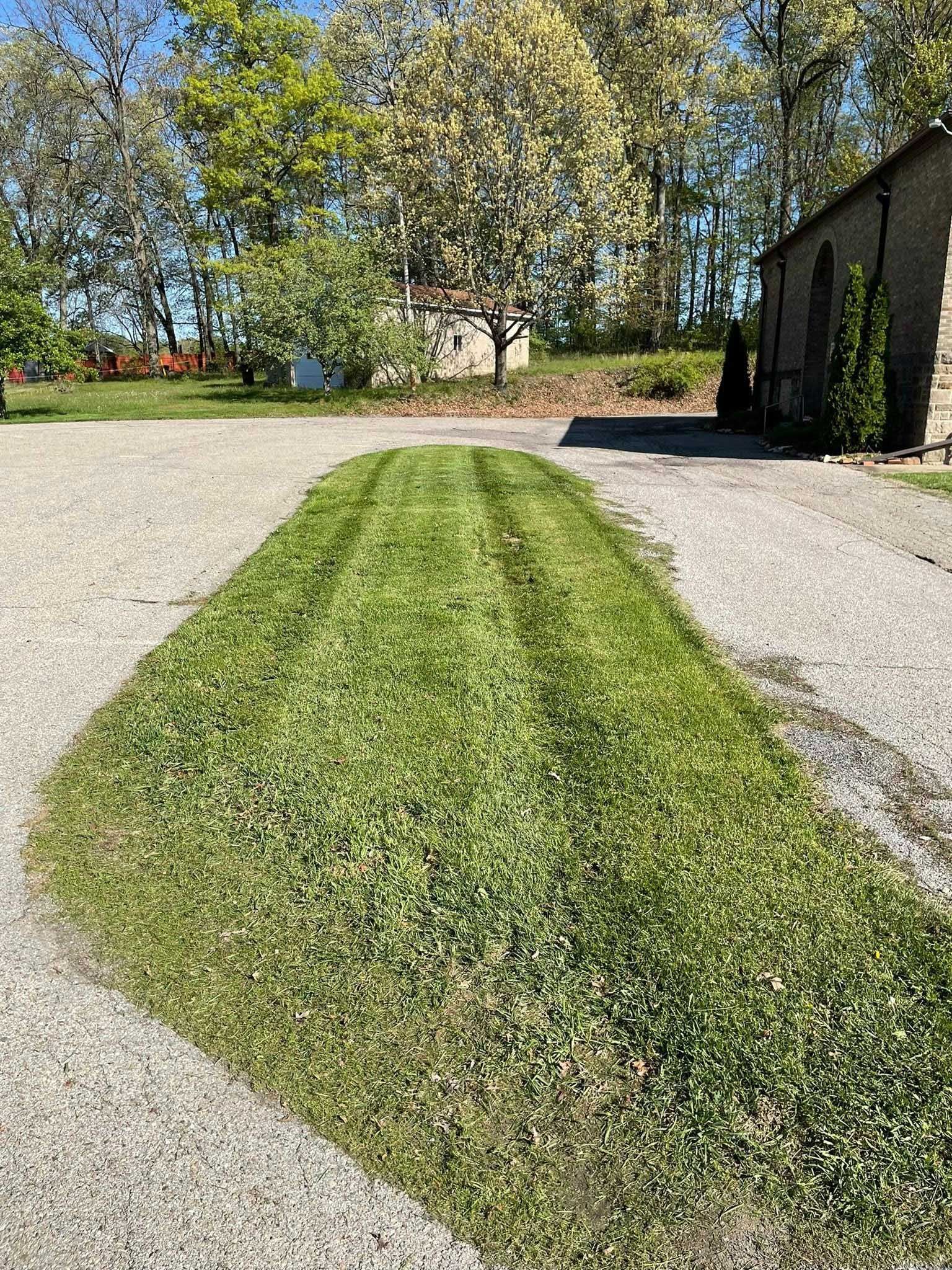 Green grass path mowed through a gravel driveway, trees and a stone building in the background on a sunny day.