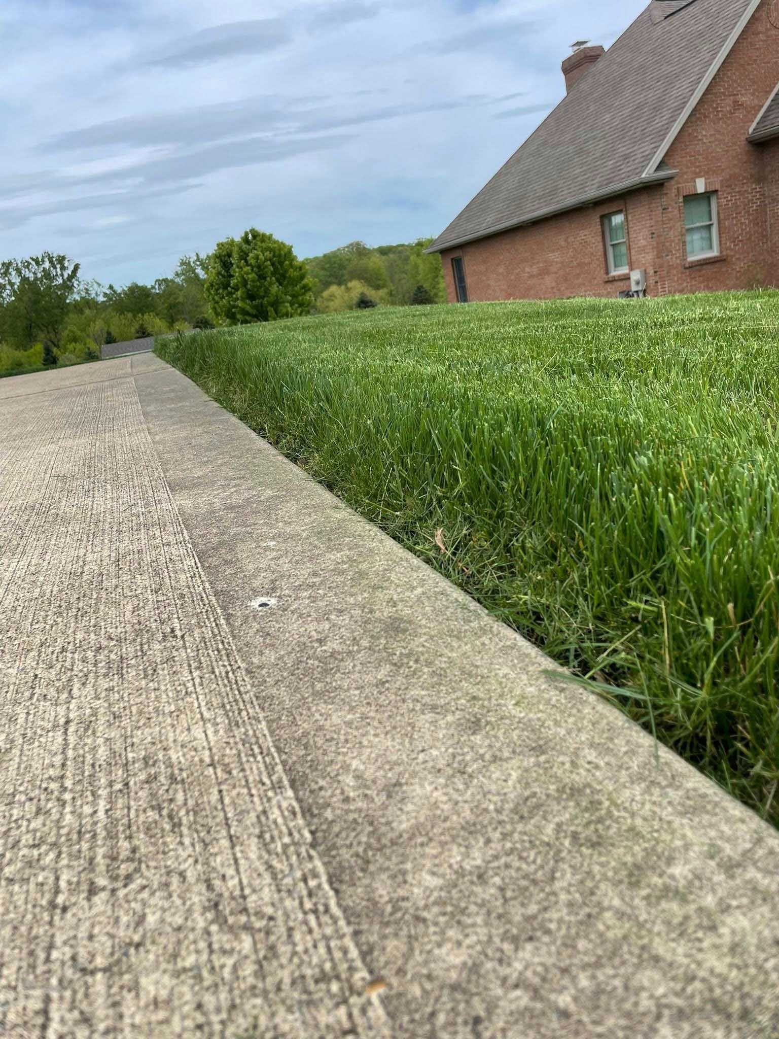 Close-up view of a concrete walkway bordering a lush green lawn next to a brick house. The sky is cloudy.