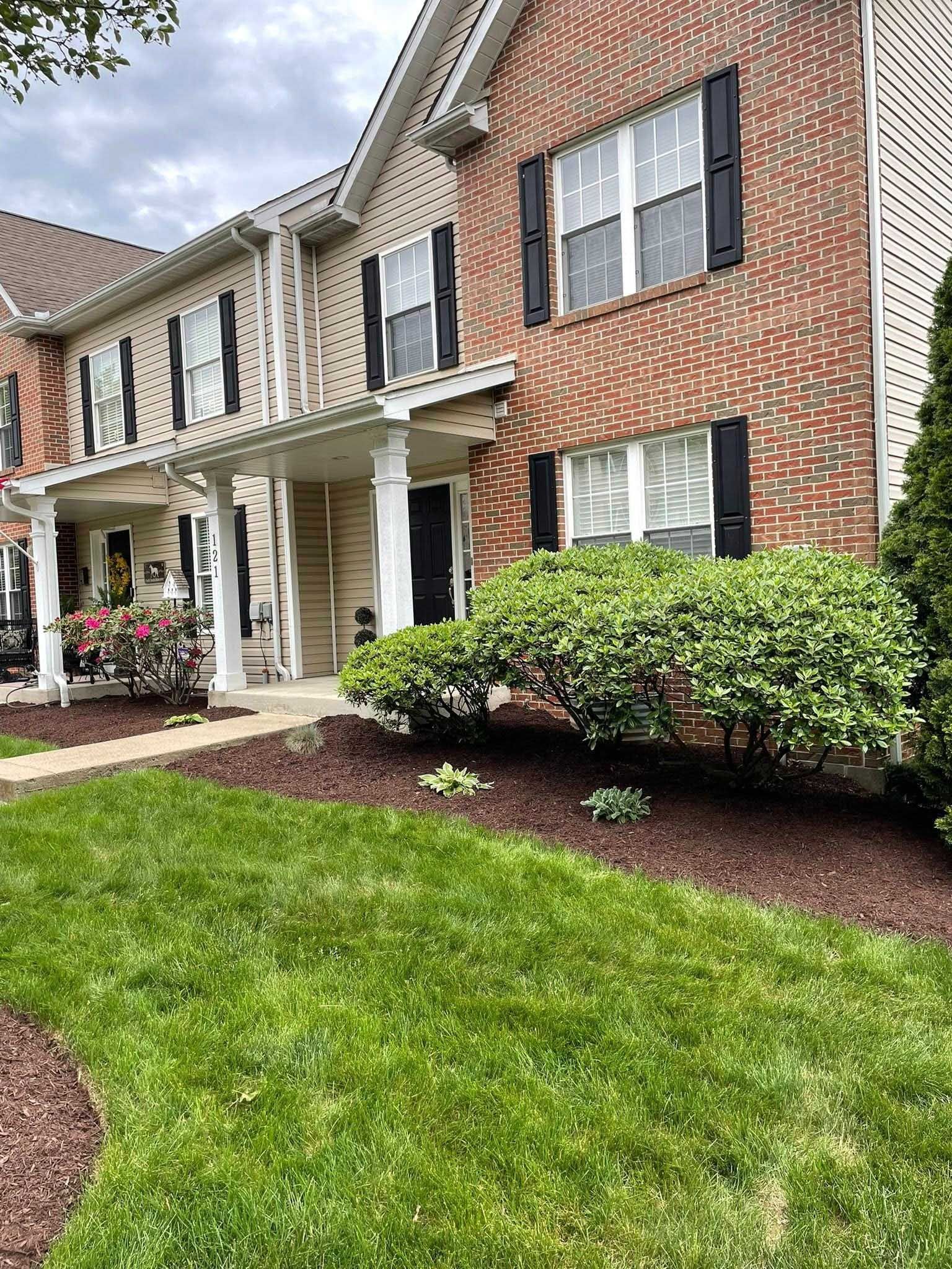 Townhouse with brick facade and black shutters, a well-manicured lawn, and a small shrub in front of the door.