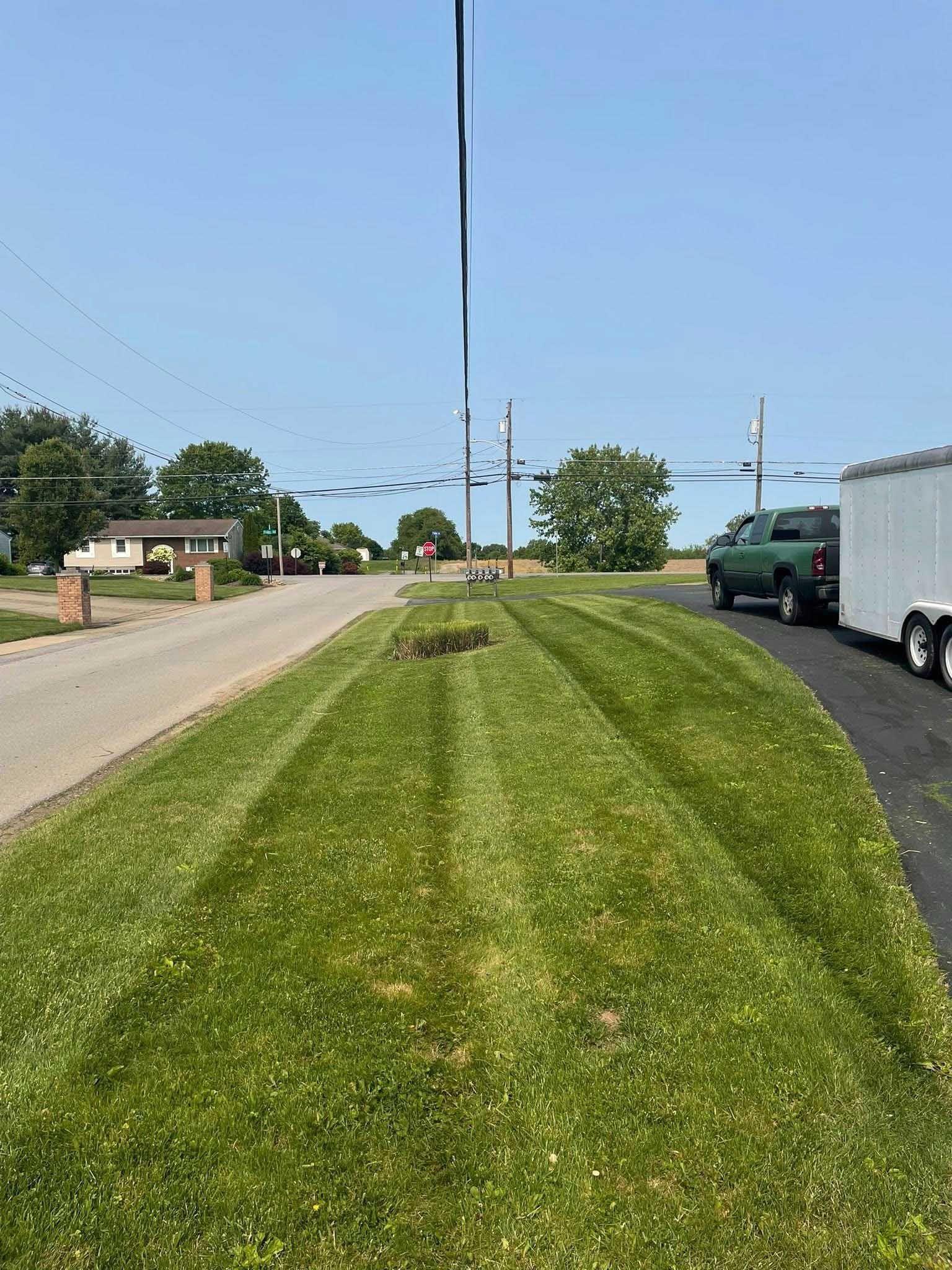 Green lawn with freshly cut stripes alongside a paved driveway, road, and parked green truck with a white trailer.