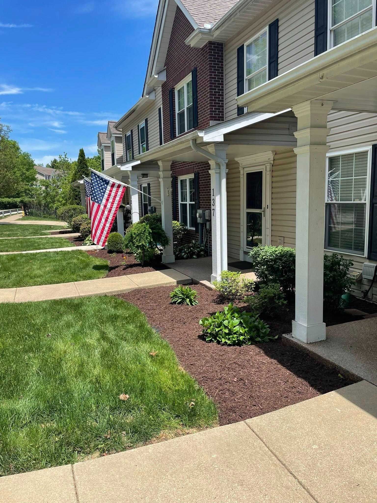 Row of townhouses with green lawns, mulch beds, and white porch pillars. An American flag hangs from one unit.