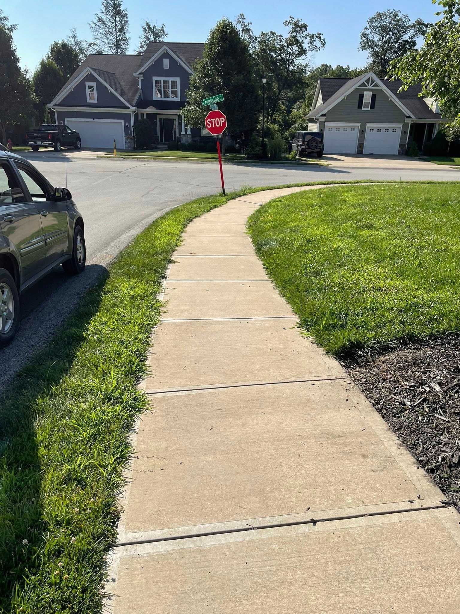 A sidewalk curves through a residential neighborhood. Lush green grass borders the concrete path. Houses are in the background.