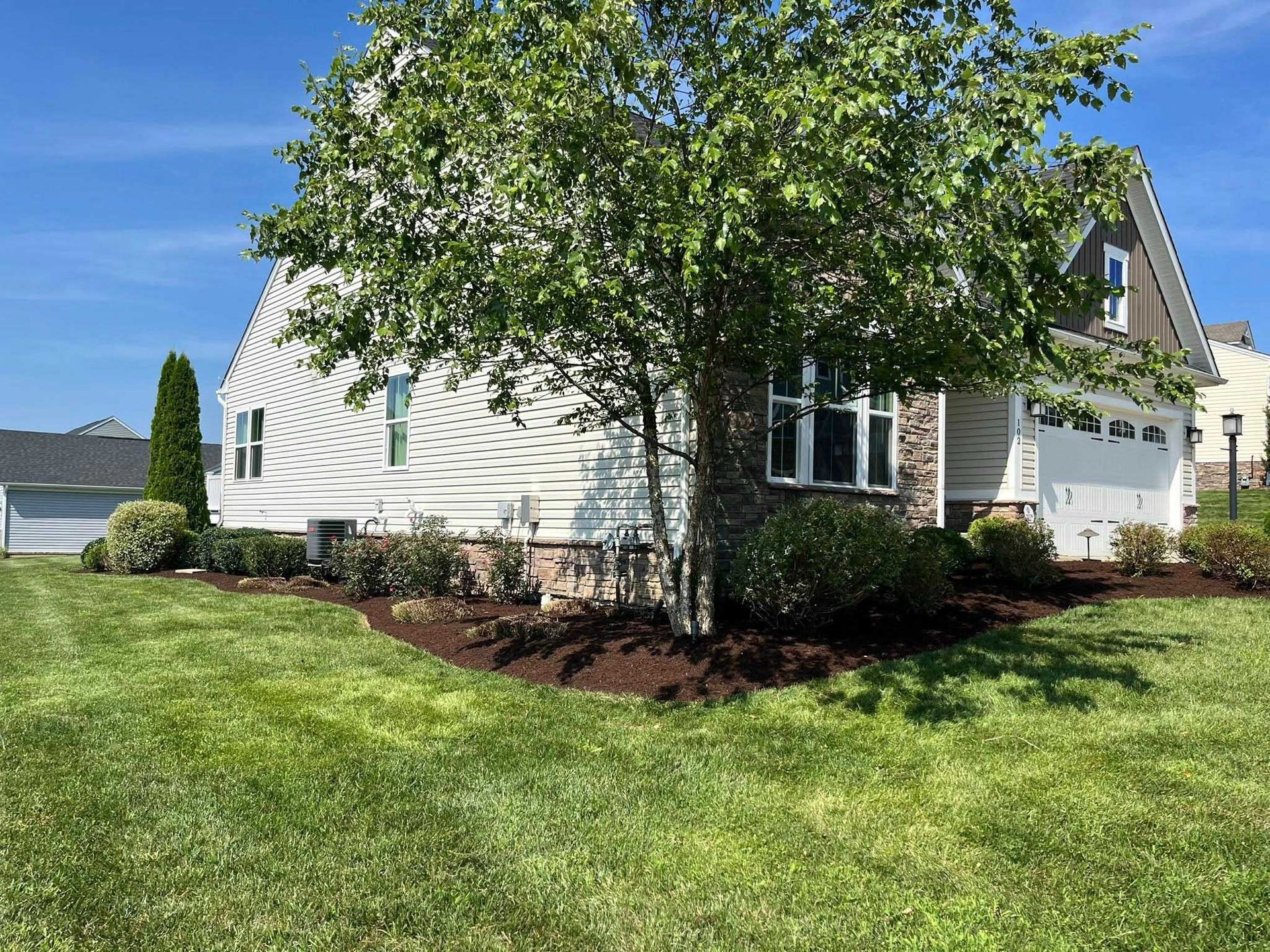 A two-story house with tan siding and a garage, surrounded by a well-manicured lawn and landscaping beds.